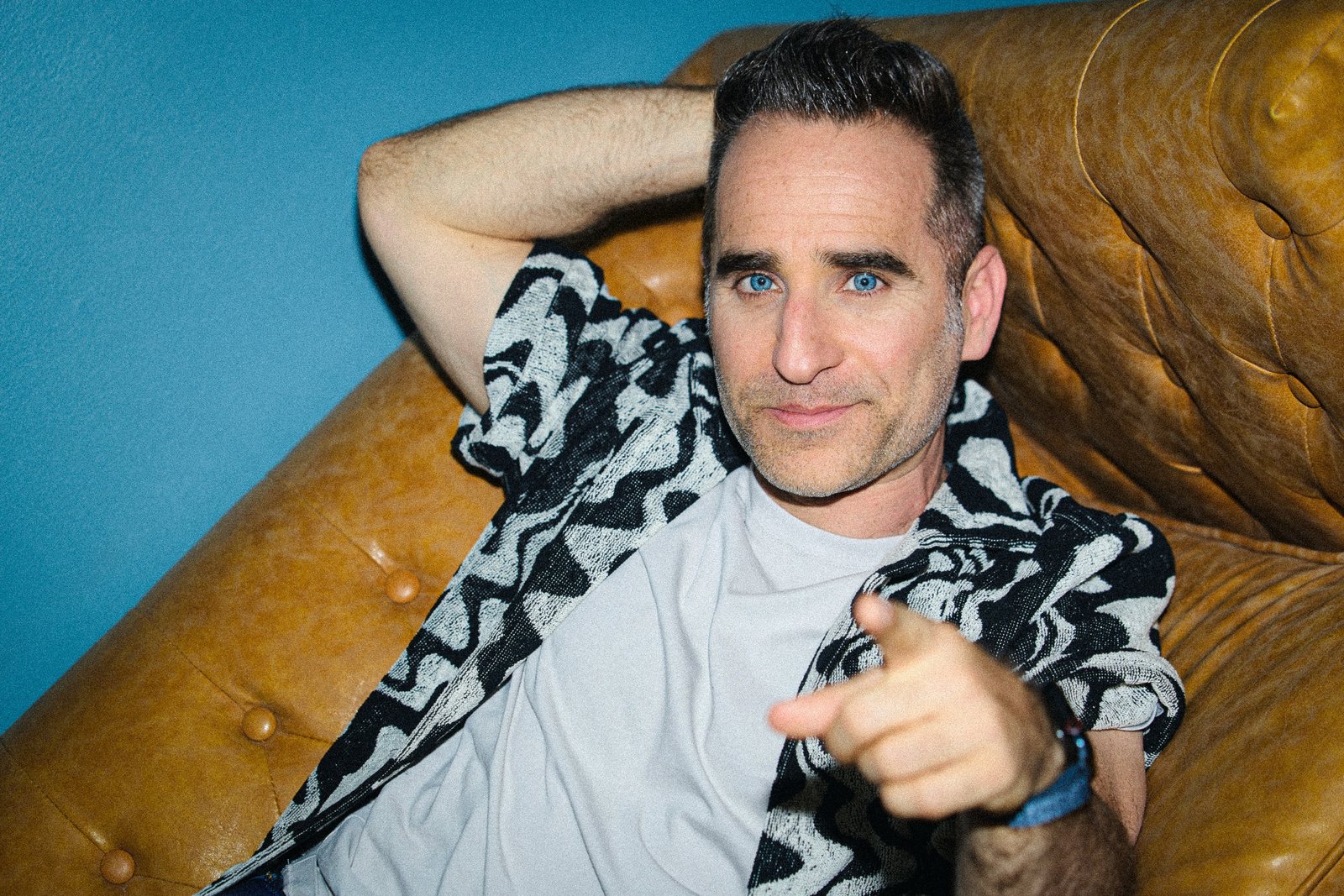 Ben lounging on a leather couch in a patterned shirt, pointing at the camera.