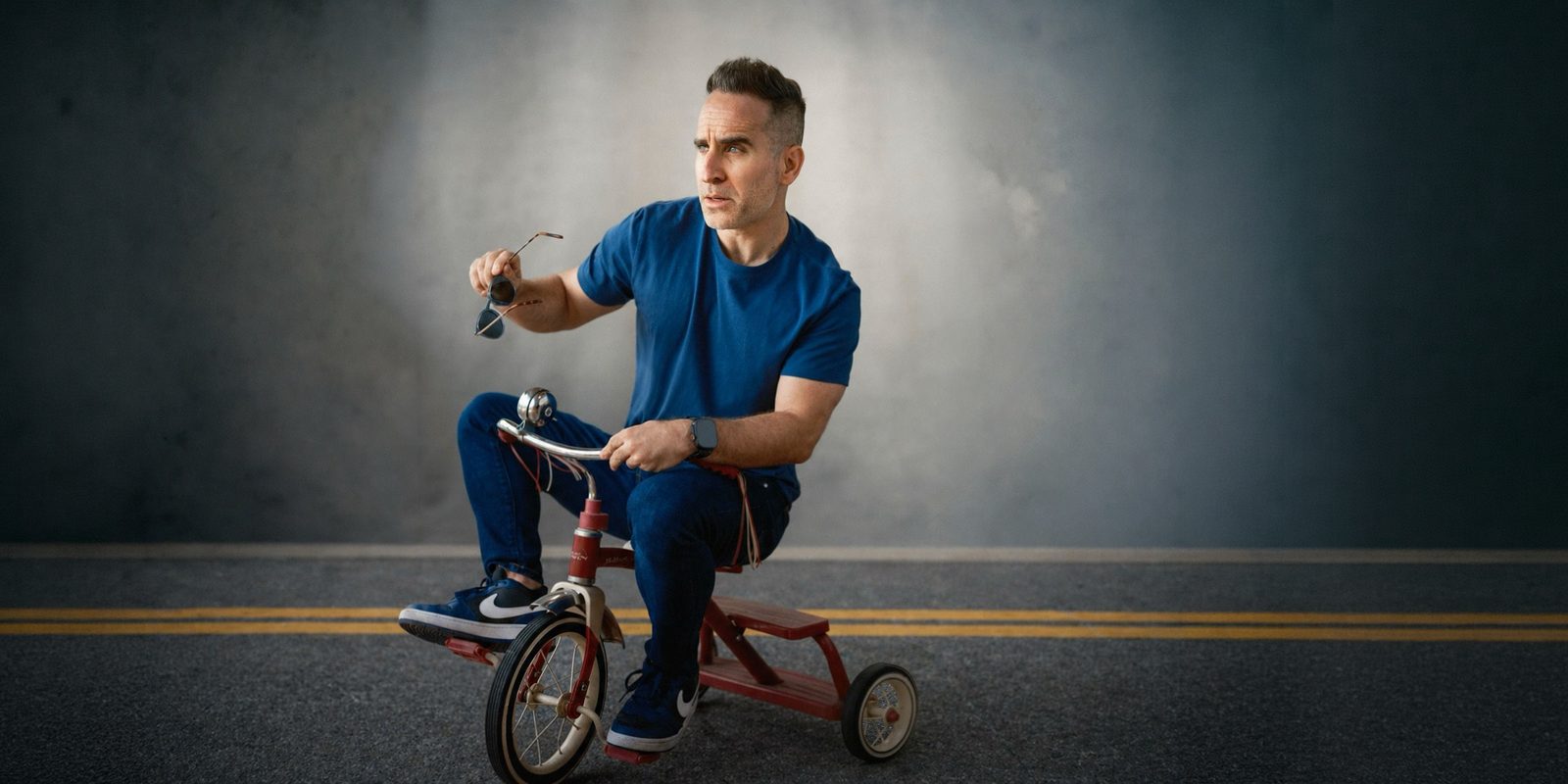 Ben Giroux on a small red tricycle in the middle of an empty road, holding his sunglasses and glaring into the distance.