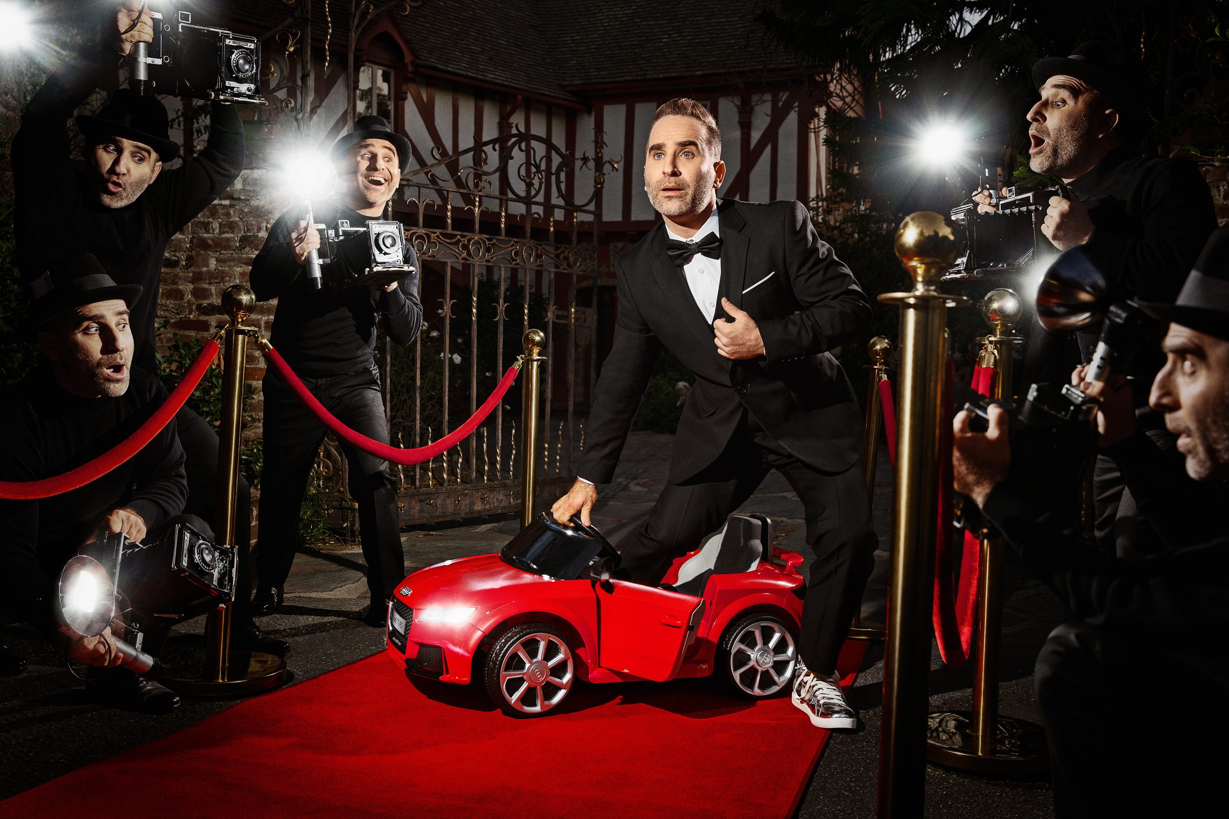 Ben in a tuxedo on a red carpet beside a tiny red toy car, surrounded by paparazzi.